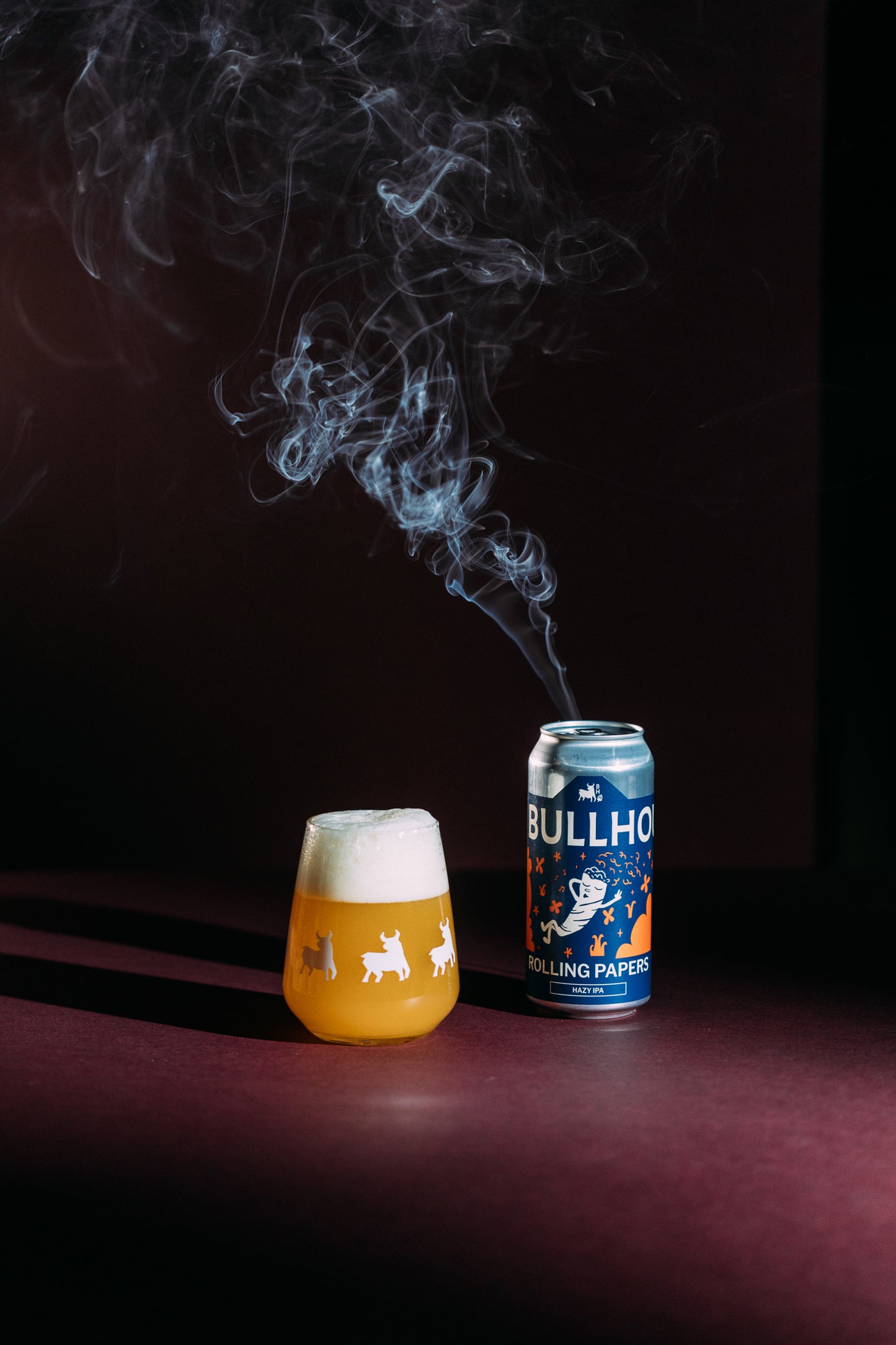 
                  
                    bullhouse rolling papers hazy ipa beer can next to a glass of beer on a red background
                  
                