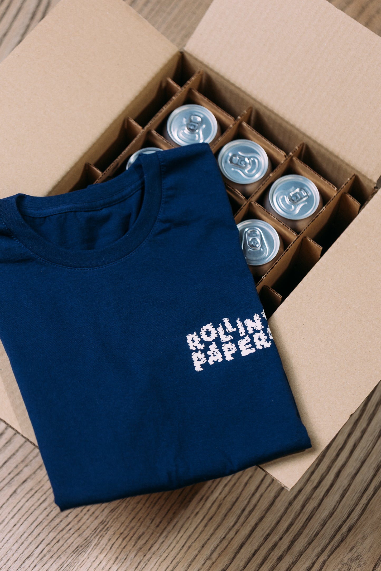 Blue t-shirt with 'Rolling Papers' text in a cardboard box with beer cans.