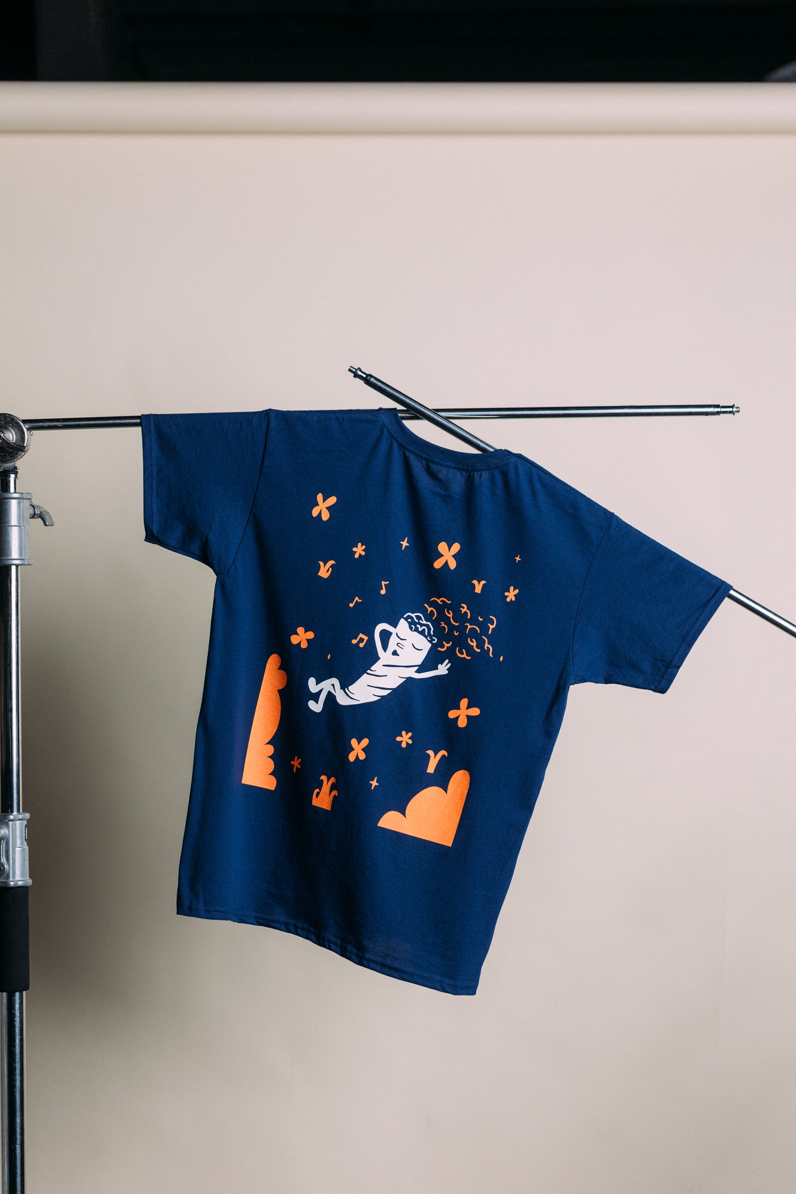 Blue t-shirt with orange graphics hanging on a rack against a beige wall.