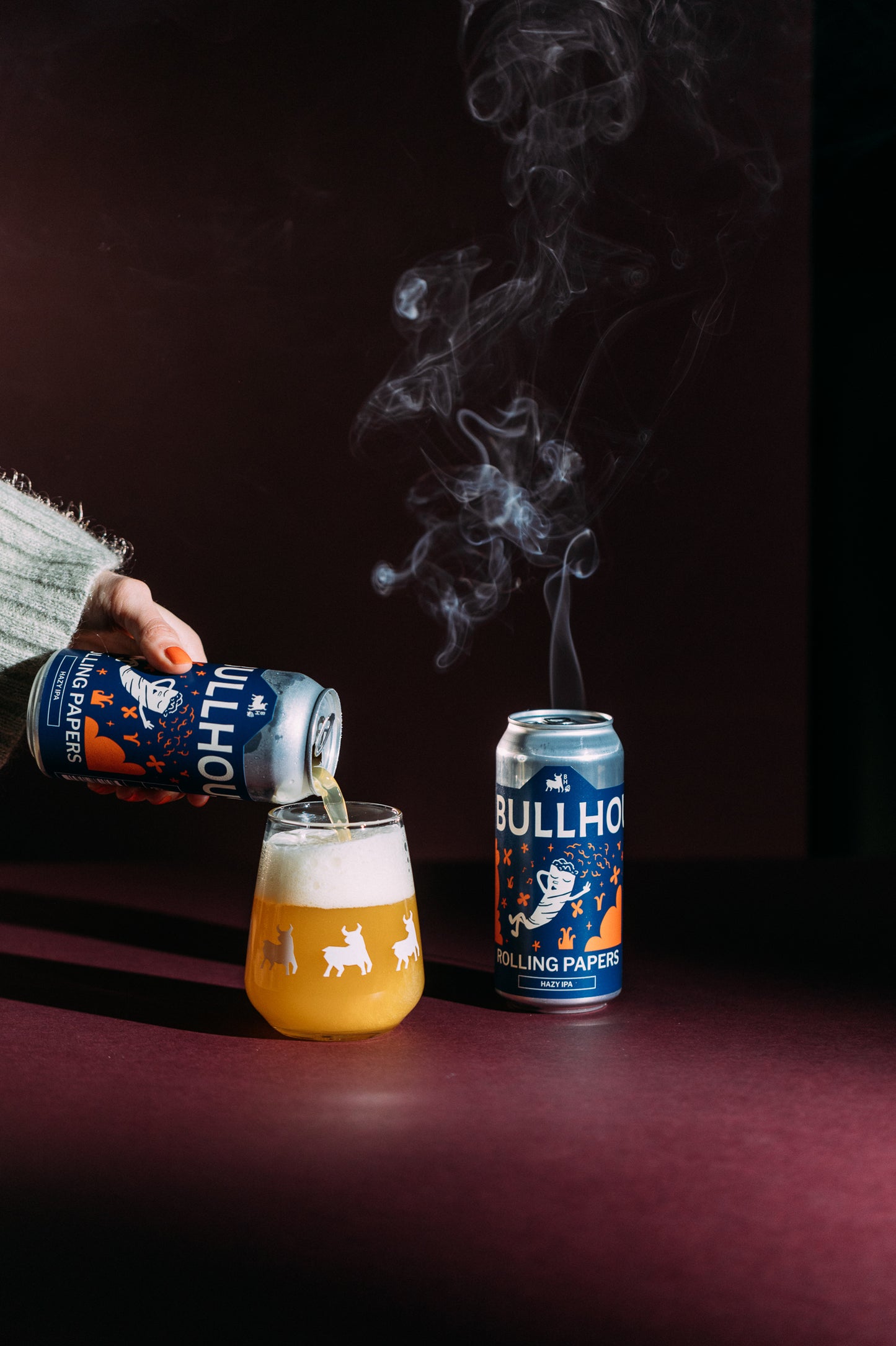 
                  
                    Person pouring a can of bullhouse rolling paper beer in a glass next to a can of bullhouse beer.
                  
                