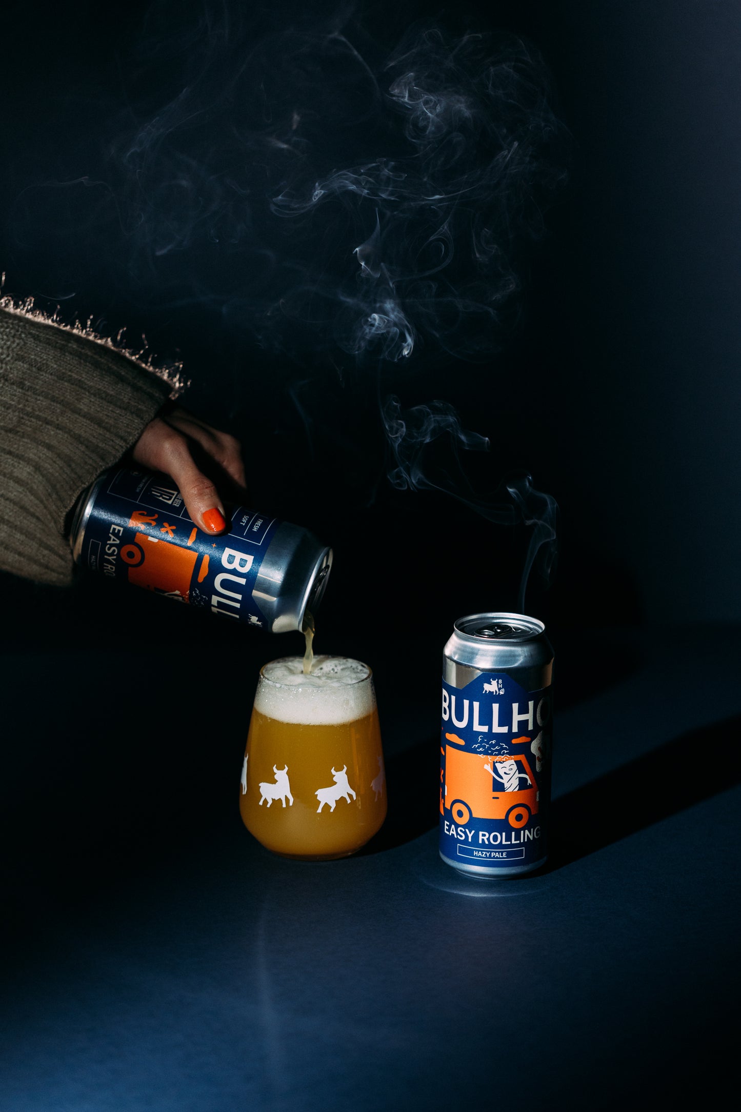 
                  
                    Person pouring beer from a can labeled 'Bullhead' into a glass with smoke effect on a dark background.
                  
                