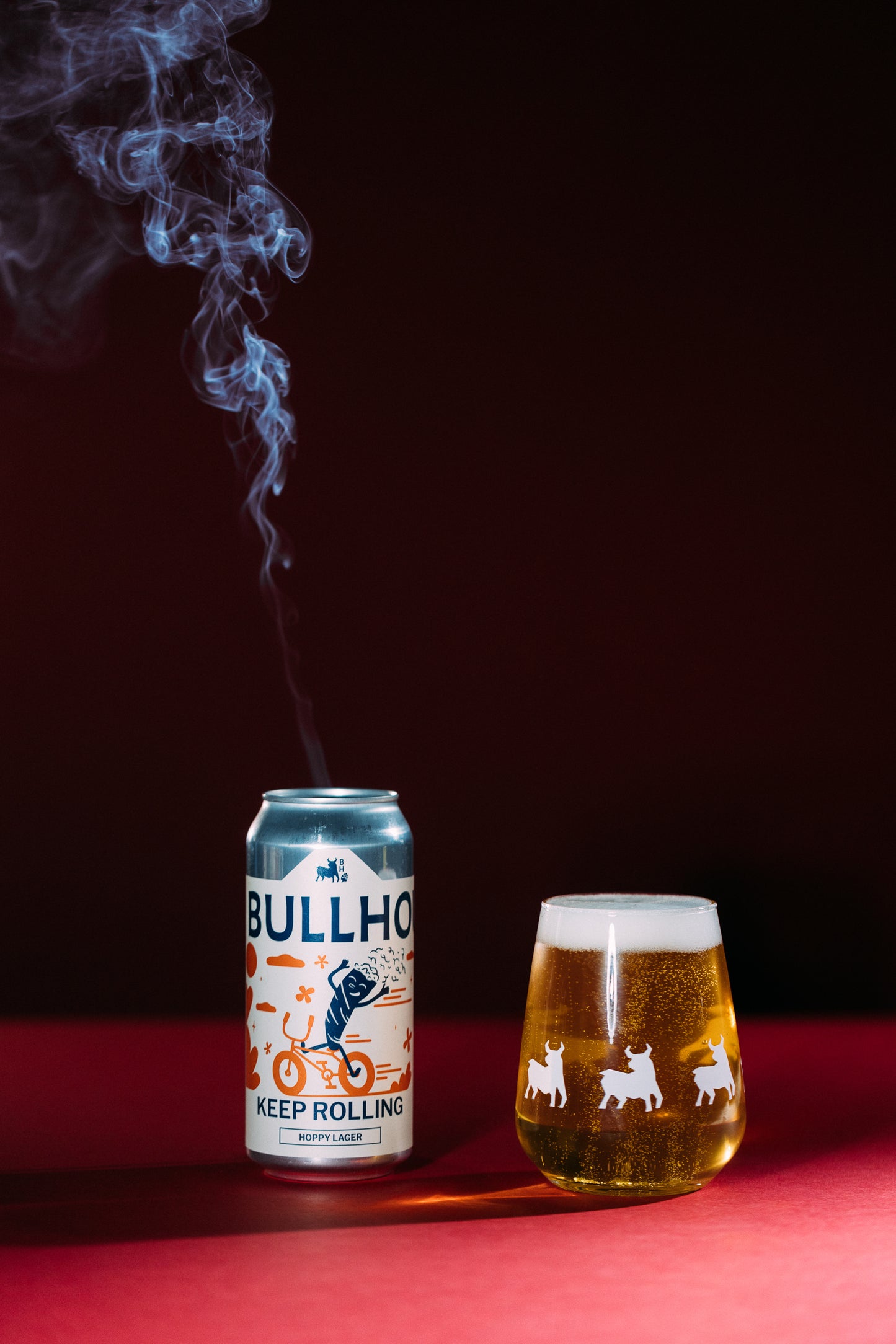 
                  
                    Bullhouse Beer can next to a glass against a red background with smoke
                  
                