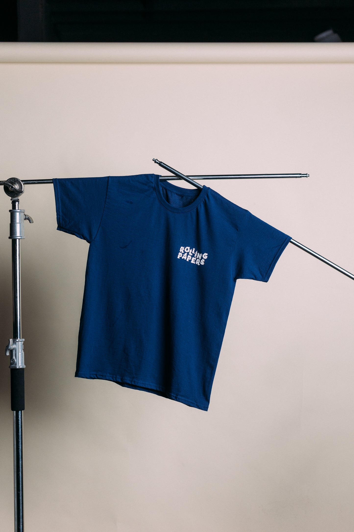 
                  
                    Blue t-shirt with a white rolling papers logo on a stand against a beige wall
                  
                